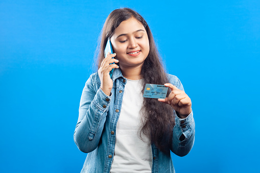 Smiling young Indian girl holding credit card and calling on cell phone