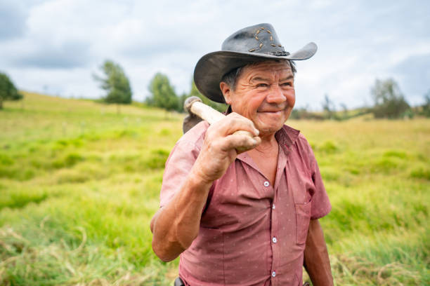 Happy Latin American senior man working at his farm and carrying a mattock on his shoulder - agriculture concepts