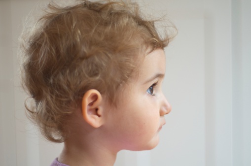Young girl, toddler, looking straight ahead, shot taken from the side. She has blonde curly hair and the background is plain. It is a close up head shot and she looks like she is looking wistfully out of a window or day dreaming.