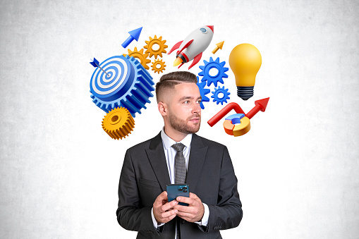 Handsome young bearded European businessman with smartphone looking sideways standing near concrete wall with startup idea icons, gears, lightbulb and target with arrow. Concept of statup goal