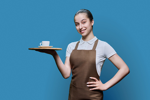 Portrait of young female waitress in an apron with tray of cup of coffee on blue studio background. Smiling girl looking at camera. Work, service in food, youth concept