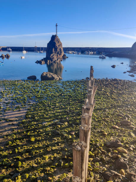 una spiaggia rocciosa con una grande formazione rocciosa sullo sfondo, un palo di legno in primo piano e barche nel mare blu profondo - beauty in nature cloud rocky coastline rock foto e immagini stock