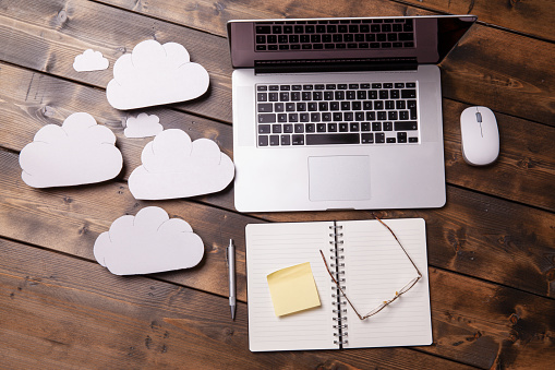 Laptop on wooden desk with cloud backup concept. Notepad and pen with glasses and mouse