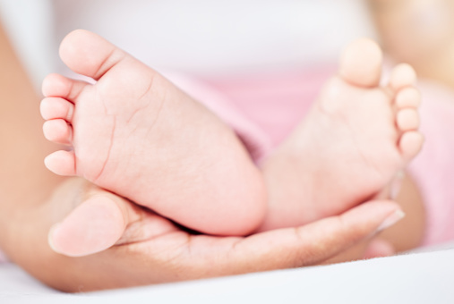 Closeup cropped view of a unrecognizable mixed race babies tiny soft feet and toes being held by the mother's hand. Woman holding the feet of her adorable newborn baby on a comfortable bed