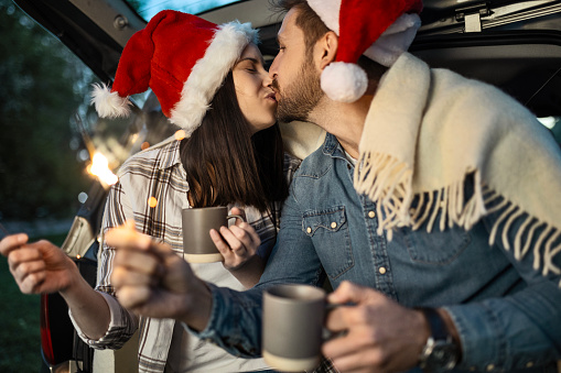 Happy couple is wearing Santa Claus hat and sitting in car trunk while enjoying moments of Christmas
