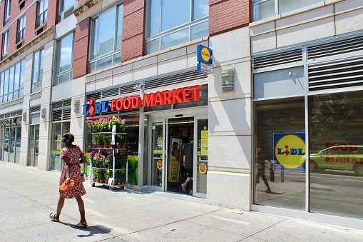 New York, NY USA - August 22, 2023 : Woman walking on the sidewalk in front of Lidl food market on Frederick Douglass Boulevard on a sunny summer day in Harlem, New York City