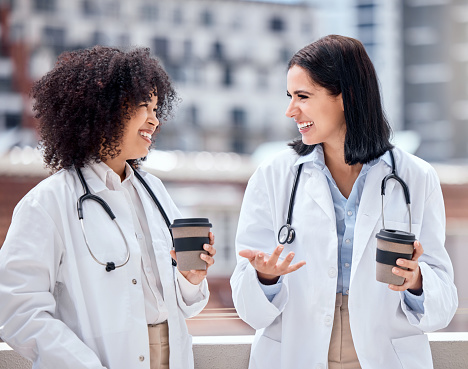 Two happy and cheerful female doctors standing and drinking a cup of coffee outside taking a break together. Caucasian and mixed race hispanic women smiling relaxing and talking on a break outside