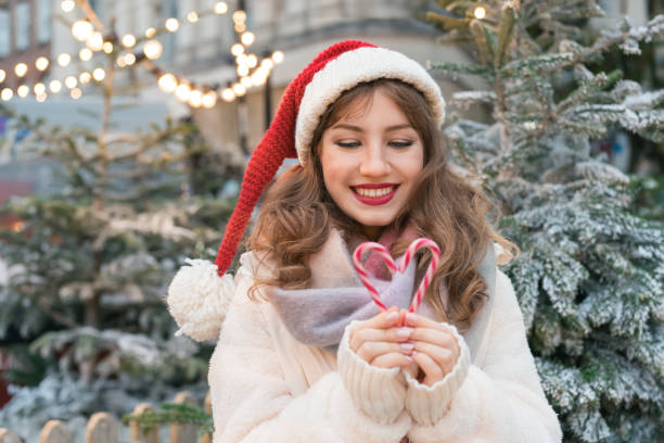 istock Young smiling woman in Santa hat making heart shape with candy canes while walking on traditional street Christmas market. Happy New Year celebration Christmas holidays. Share love, charity concept 1816811745