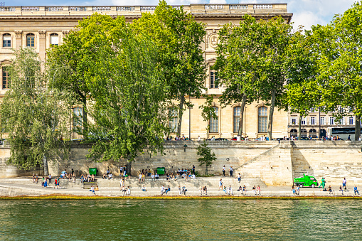 Parisians and tourists sitting and walking on the quays of the Seine river on a sunny summer day in Paris, France