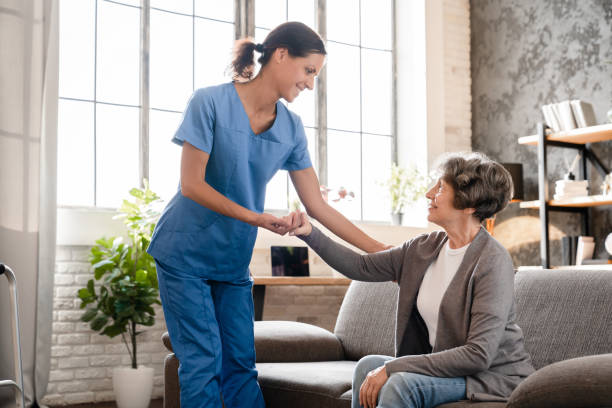 istock Nurse caretaker caregiver doctor helping assisting aiding old elderly disable immobile handicapped patient grandmother stand up from the couch. Rehabilitation after injury trauma 1816839129