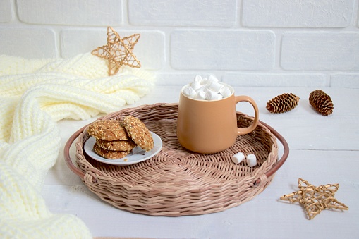 Brown mug with hot drink and marshmallows, a saucer with cookies on round wicker tray. Cozy Christmas breakfast or snack.