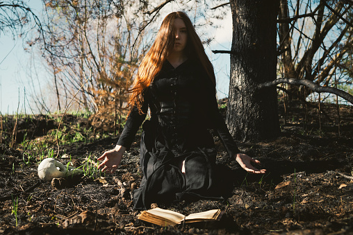 Mystical witch red haired woman in a black long dress in a gloomy sitting forest with book. Gothic girl on a scorched ground.