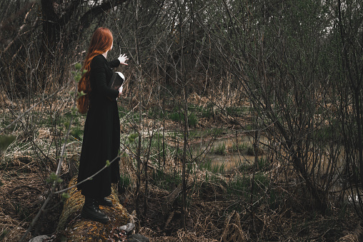 Gothic red haired woman in a black long dress in a gloomy forest with book. Mystical witch girl.