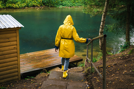 Woman in a yellow raincoat and rubber boots descends the stairs to the pier by the lake in a green wet forest during the rain. The concept of travel and tourism.