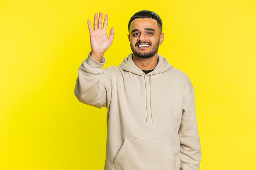 Young Indian man smiling friendly at camera, waving hands gesturing hello greeting or goodbye welcoming with invitation hospitable expression. Arabian Hindu guy isolated on yellow studio background