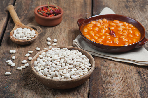 High angle photo of a bowl of navy beans on a vintage wooden table with traditional home cooked navy beans dish on a linen napkin in the background