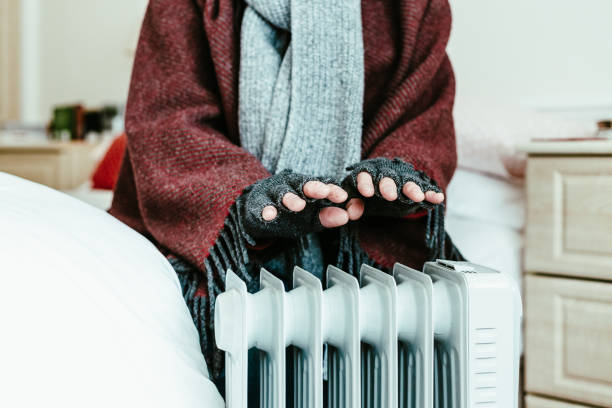 Cold senior man warming his hands over electric heater at home
