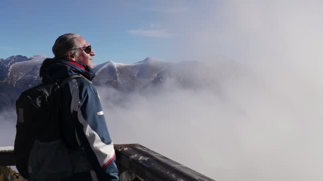 A gray-haired man with sunglasses stands high in the mountains against the backdrop of a mountain range and watches the moving clouds