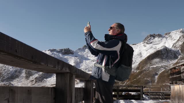 A gray-haired adult man stands against the backdrop of high mountains and films a video on his mobile phone.