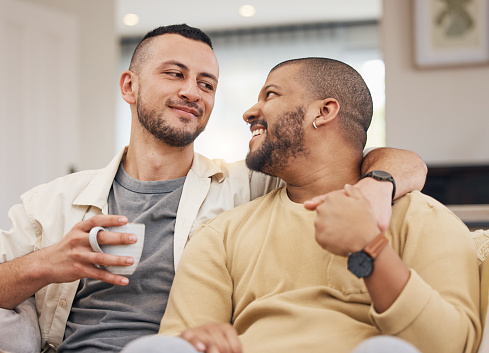Happy, love and gay couple relaxing on a sofa with a cup of coffee in the living room together. Smile, bonding and young lgbtq men with a latte sitting in the living room of their modern apartment.