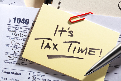 A yellow note pad rests on top of a 1040 tax form with a reminder that it's tax time.
