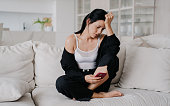 Sad brunette American woman sitting on couch reading message looks at phone, frustrated after receives awful news. Unhappy woman touching head, upset, thinks about future. Failure, crisis.