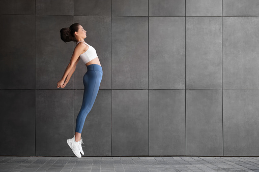 Beautiful woman in sportswear jumping near dark grey wall outdoors. Space for text