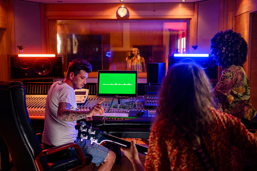 Photo of a vocal recording session in a music studio. The music producer lifter his arm to ask for silence of other band mates while the singer is singing. Focused, attentive, listening carefully.