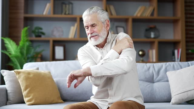 Senior gray haired bearded man suffers from sharp pain in his shoulder while sitting on sofa in living room at home.