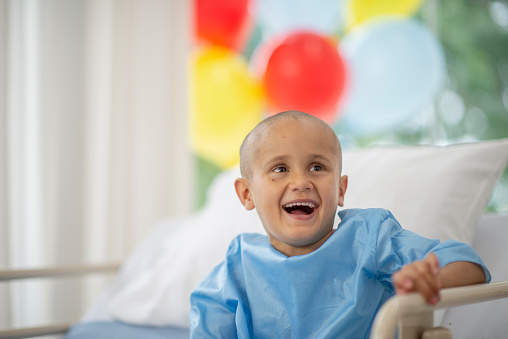 A sweet little boy with cancer, sits up in his hospital bed with a big optimistic smile on his face as he prepares to have another treatment.  He is wearing a blue hospital gown, has his head shaved.