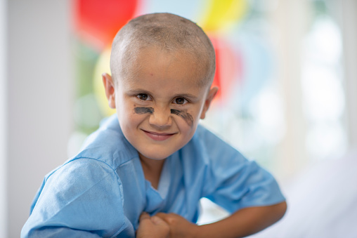 A mixed race oncology patient sits up in his hospital bed, in his bright blue gown, with warrior paint under each eye.  He has a strong and serious expression on his face as he flexes his muscles.