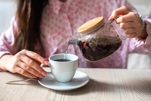 woman female hands hold transparent teapot with herbal healthy warming tea and pour hot tea into white cup