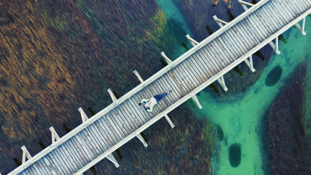 AERIAL Drone Zoom Out Shot of Female Tourist Lying on Wooden Footbridge Over Marshy Turquoise River in Zelenci Nature Reserve
