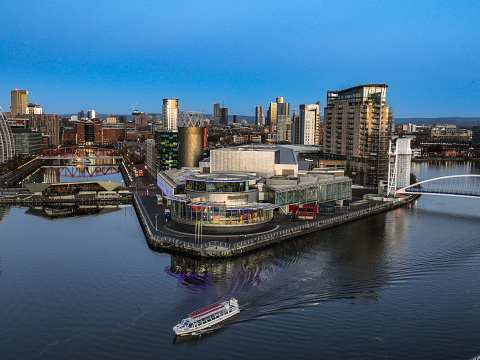 An aerial photograph of Salford Quays at dusk. The photograph was produced in early winter as the sun was setting. The photograph shows a tourist boat travelling along the Manchester Ship Canal. Downtown Manchester can be seen in the distance.