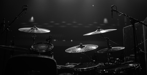 A drum set sits on a dimly lit stage, its cymbals and drums gleaming in the spotlight. The stark contrast of the black and white tones emphasizes the intricate details of the drum set, from the grooves on the cymbals to the texture of the drumheads. The overall effect is one of anticipation and excitement as if the drum set is waiting to be played.
