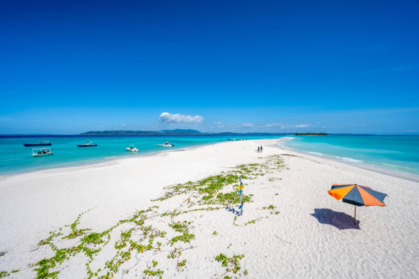 Paradise-like, white sandy beach with sunshades, tourists, leading to a beautiful island and leading into the turquoise blue, clear sea on both sides. Nosy Be, Nosy Iranja, Madagascar, Africa. Description: Paradise-like, white sandy beach with sunshades, tourists, leading to a beautiful island and leading into the turquoise blue, clear sea on both sides. Nosy Be, Nosy Iranja, Madagascar, Africa. madagascar beach photos stock pictures, royalty-free photos & images