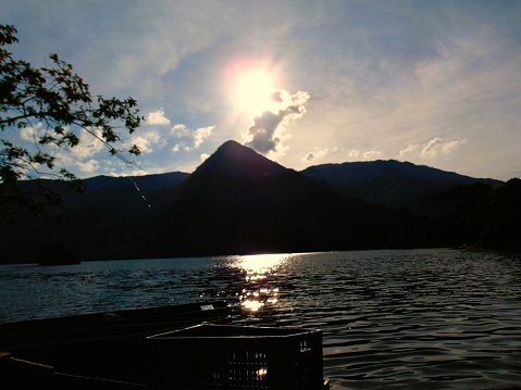 Lagoon between forests and mountains, sun, clouds, reflection of the sun, boats, water, reflection of the blue sun.