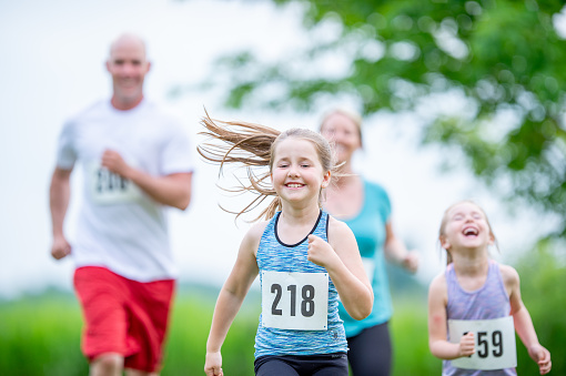 A small family of four are seen running together in a cross country race on a beautiful summer day.  They are each dressed comfortably in athletic wear and are wearing numbered bibs as they focus on their course.