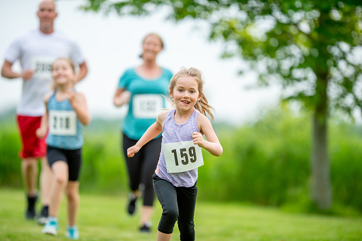 A small family of four are seen running together in a cross country race on a beautiful summer day.  They are each dressed comfortably in athletic wear and are wearing numbered bibs as they focus on their course.
