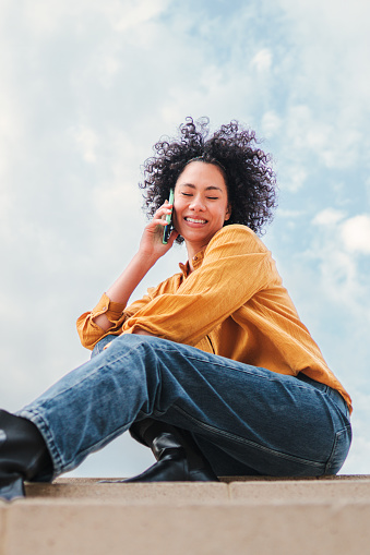 Vertical portrait of young adult hispanic woman with curly hair having a cellphone call conversation sitting under blue sky with clouds. Happy latin girl talking by phone, at background the skyline.