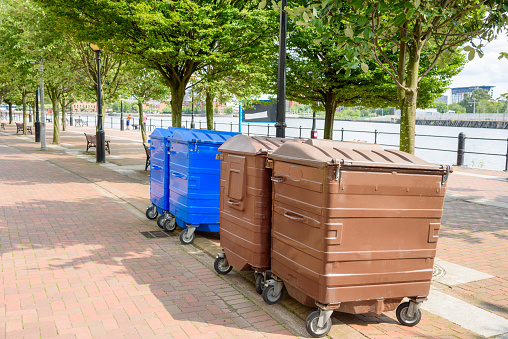 Large recycling wheelie bins along a tree lined harbourside footpath on a sunny summer day. Salford Quays, Manchester, England, UK.