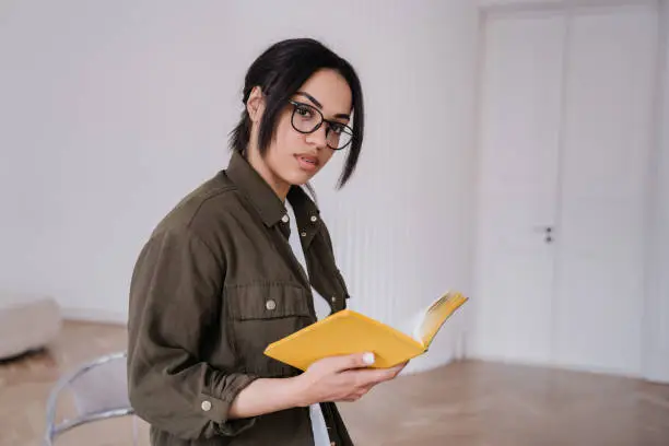 Puzzled young woman in glasses and casual shirt standing at home holds notebook looks away with thoughtful face expression. Lonely girl reading poetry at home. Hesitating female indoors.