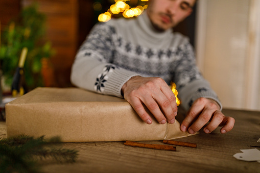 Selective focus shot of neat young man folding edges of a rustic brown paper that he is using to wrap a Christmas gift box.
