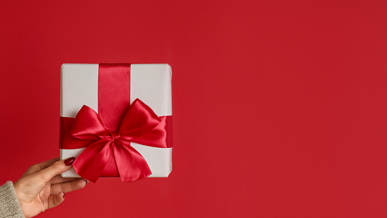 Christmas present. Winter holiday surprise. Female hands showing white gift box with bright ribbon bow isolated on red empty space background.