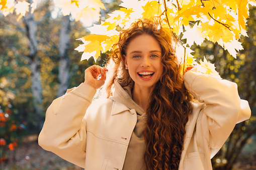 Smiling young woman enjoys the autumn weather above the swirling fall foliage tree in the forest with the yellow leaves at sunset