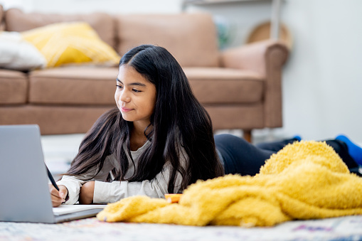 A young adolescent of Hispanic decent, lays on her living room floor with her laptop open in front of her as she attends school virtually.  She is dressed casually and taking notes.