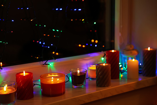 Many burning candles and festive lights on window sill indoors. Cosy atmosphere