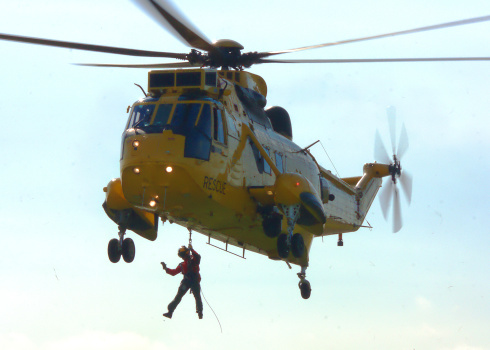 A crew member from a hovering military rescue helicopter is lowered by powered winch from a side door.
