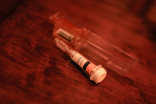 A close-up shot of a prefilled hypodermic syringe that has already been used. The plunger has been completely pressed down and is locked in the used position, with the needle shield fully extended to prevent any injury or accident. The injection was prescribed to prevent post-surgical blood clots. The packaging it came in, is placed on the wooden table behind the used syringe. Focus on the barrel. Horizontal format. No people. Copy space. Note to Inspector: Logos, company name and batch numbers have been removed. Only part of the expiry date, country of manufacture and an instruction that does not make much sense remain.