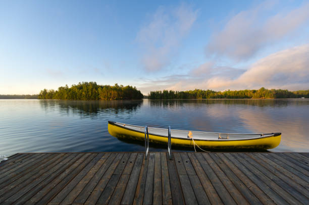 A yellow canoe is securely fastened to a rustic wooden dock nestled in the serene beauty of Muskoka, Ontario, Canada. In the distance, quaint cottages dot the landscape across the tranquil lake.